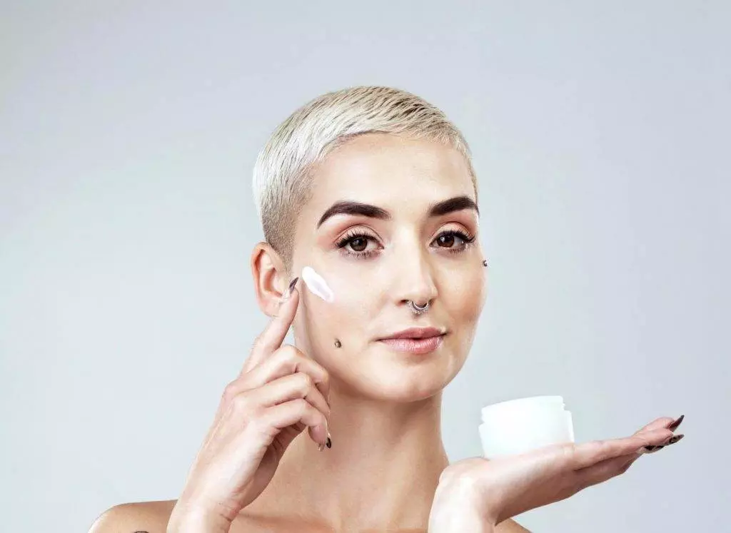 A woman with a shaved head is applying a touch-up cream to her face.