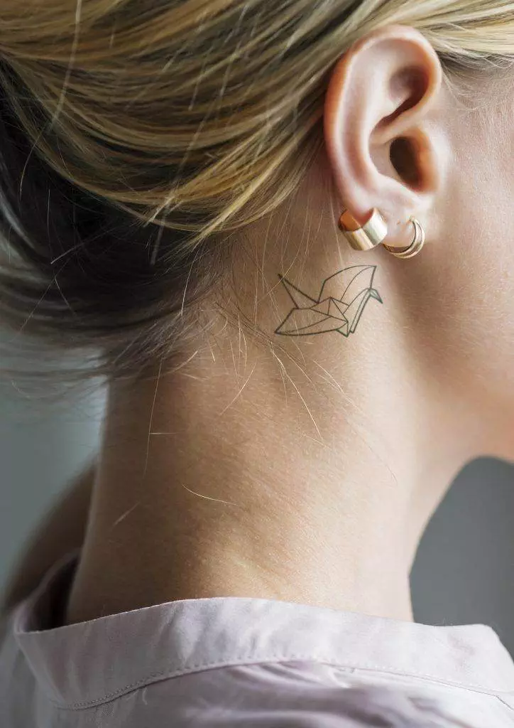 Close-up of a woman's neck in San Diego, featuring a minimalistic origami bird tattoo behind the ear. She wears two gold hoop earrings.