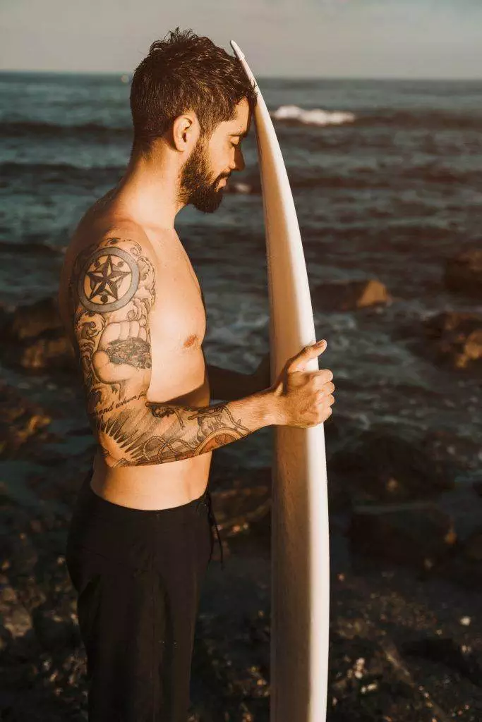 A man with San Diego tattoos stands shirtless on a rocky shore, holding a surfboard vertically, framed by the vast sea in the background.