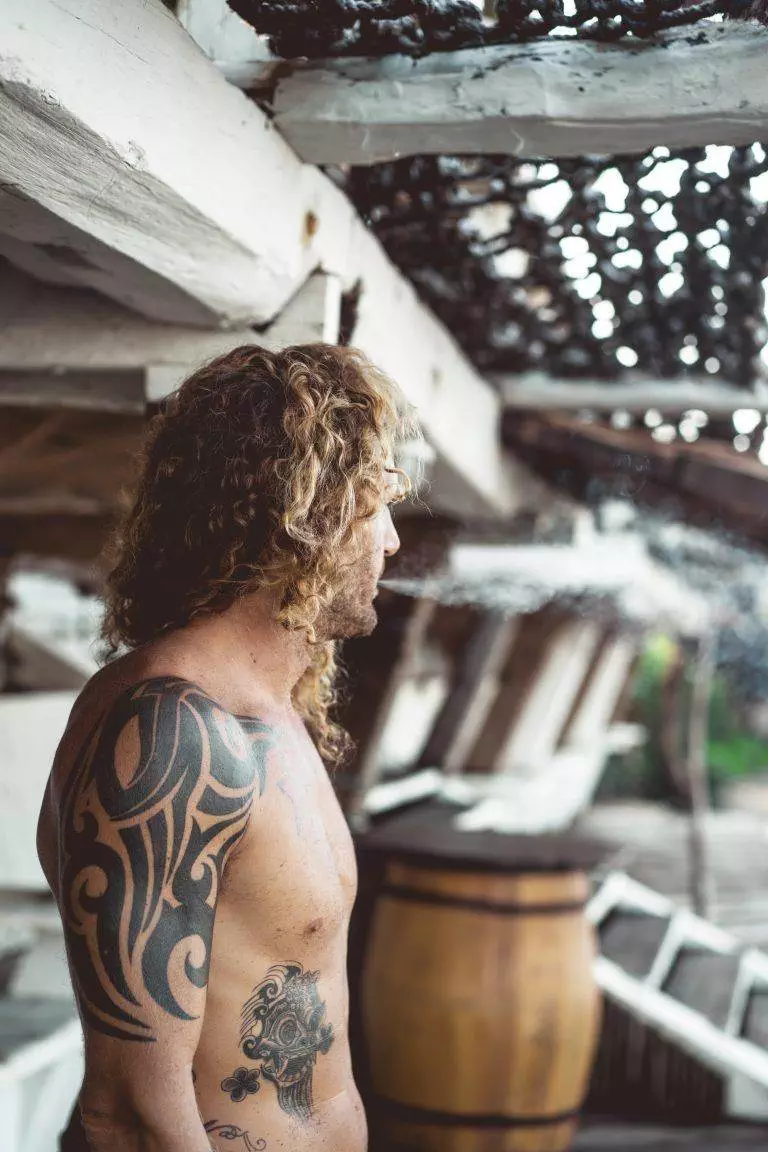 A man with curly hair and a Funhouse Tattoo tribal tattoo on his shoulder looks away from the camera, standing by a wooden structure.