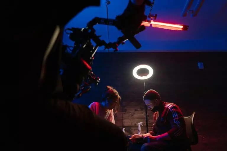 Two people seated under a ring light, one operating equipment, in a dimly lit room with blue and red lighting, reminiscent of the meticulous cleanliness found in top-tier tattoo studios.