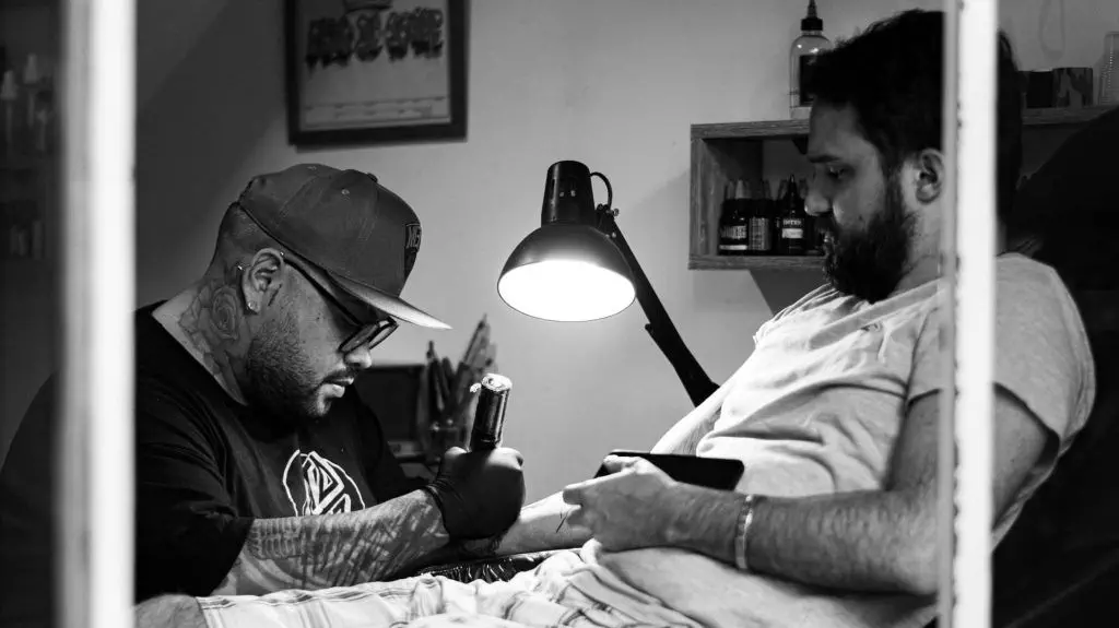 A tattoo artist with a cap and gloves works on a client's arm in a small studio, both focused, under the lamp illuminating the workspace—highlighting the importance of cleanliness in tattoo studios.