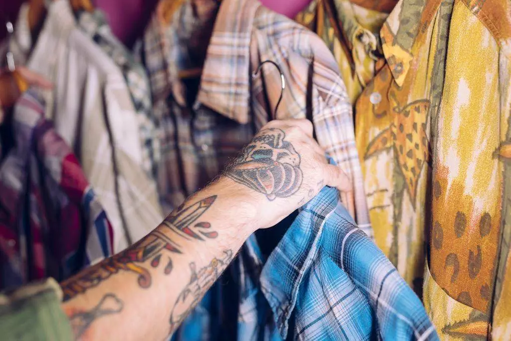 A San Diego tattoo peeks out as an inked arm reaches for a plaid shirt amidst a collection of patterned garments hanging on a clothing rack.