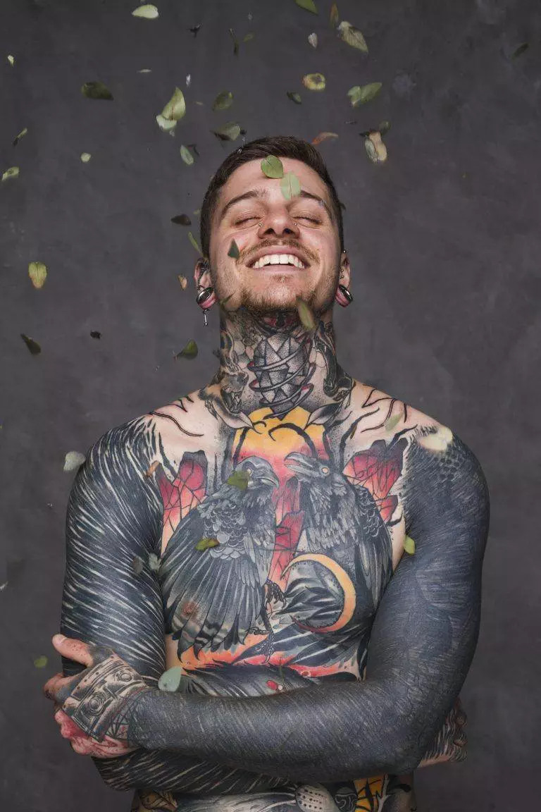 A person with expansive San Diego tattoos, featuring birds and geometric designs, smiles with eyes closed as leaves fall around them against a gray background.