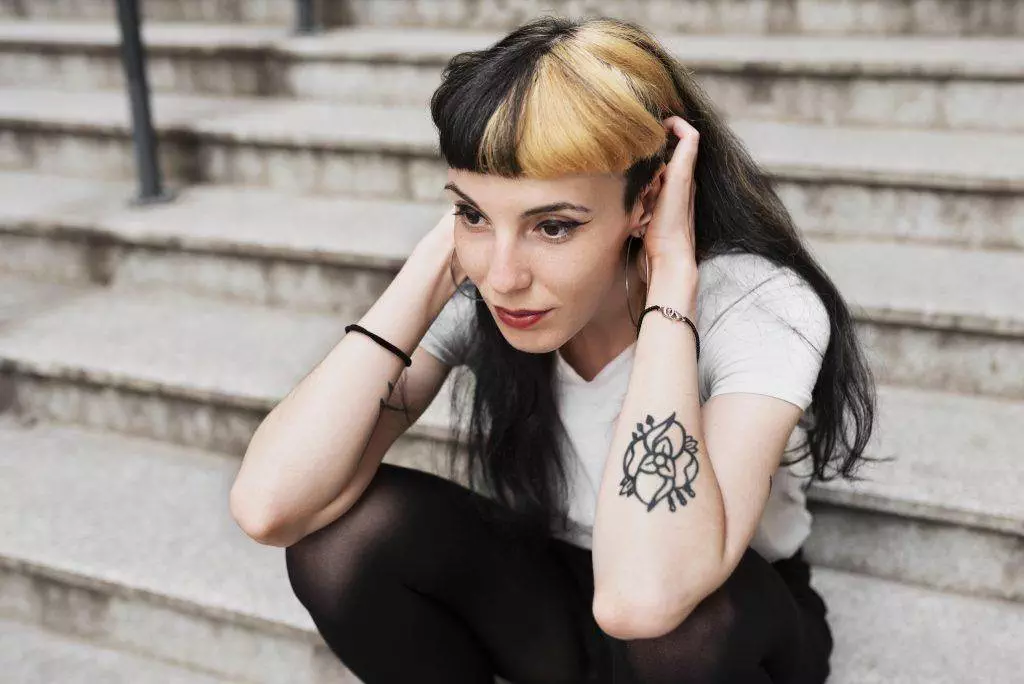 A woman with black and blonde hair sits on the steps at Pacific Beach, holding her head with both hands. She is wearing a white t-shirt, black pants, and showcases a New School tattoo of a rose on her left forearm from Funhouse Tattoo.