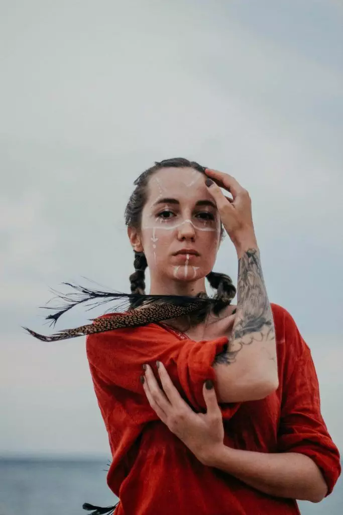 A person with braided hair, facial paint, and a tribal tattoo on their arm stands outdoors, wearing a red shirt and holding a feathered item near their shoulder, each element reflecting the cultural significance of stories behind the ink.