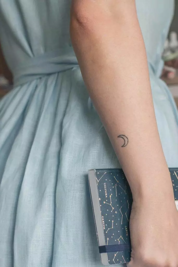 A person in a light blue dress holds a book with a constellation-covered cover in their right hand. Their left arm, adorned with a small crescent moon tattoo—perhaps an inspiration for the tattoo industry—is visible, a visual detail perfect for sharing on social media.