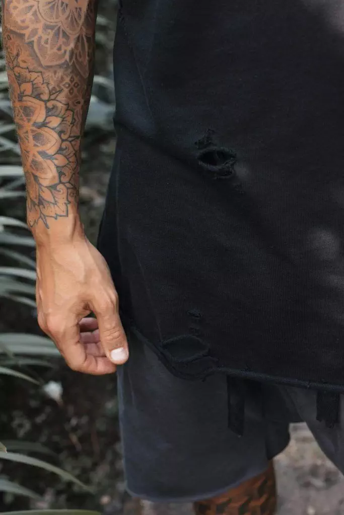 A person with tattooed arms is wearing a black, torn t-shirt and dark shorts, showcasing the latest trends in the tattoo industry.