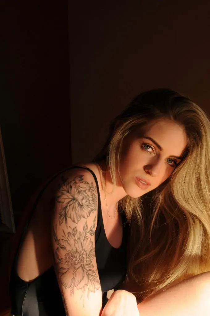 A woman with long blonde hair, wearing a sleeveless top, reveals a floral tattoo on her left upper arm. In a dimly lit setting, she gazes directly at the camera, embodying the edgy aesthetics celebrated in the tattoo industry and often showcased on social media.