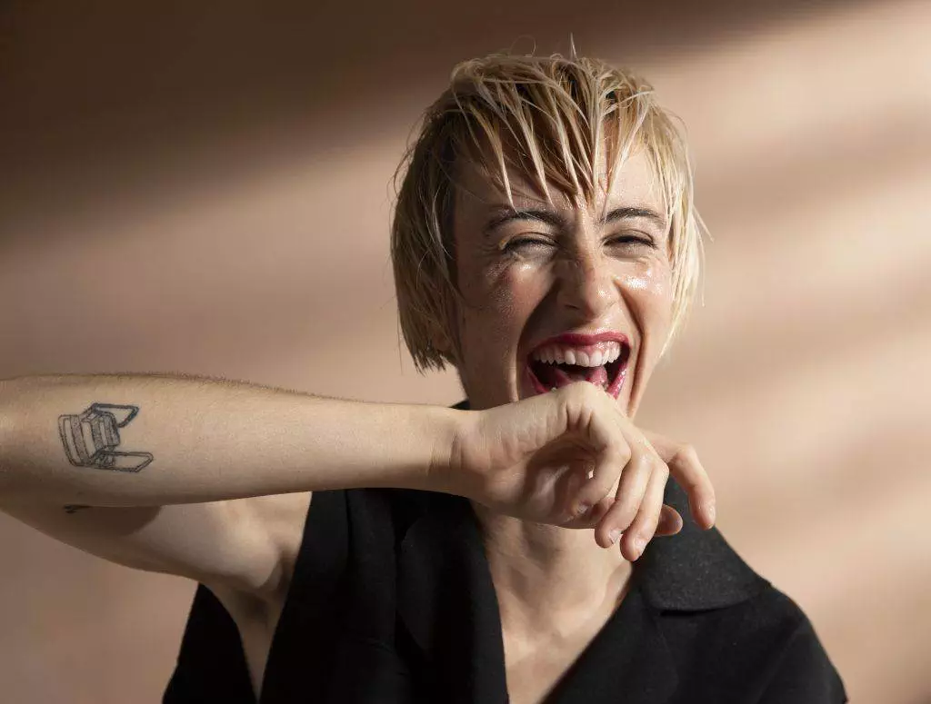 Person with short, wet hair and a visible Fine Line Tattoo on their arm smiles broadly while covering part of their mouth with their arm. Blurred, light brown background.