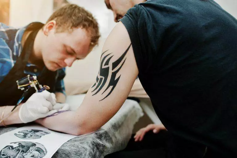 A tattoo artist with gloves is applying a new New School Tattoo on a person's upper arm, which already features a large, black tribal-style design. The artist is focused, and tattoo reference sheets from Funhouse Tattoo in Pacific Beach are nearby.