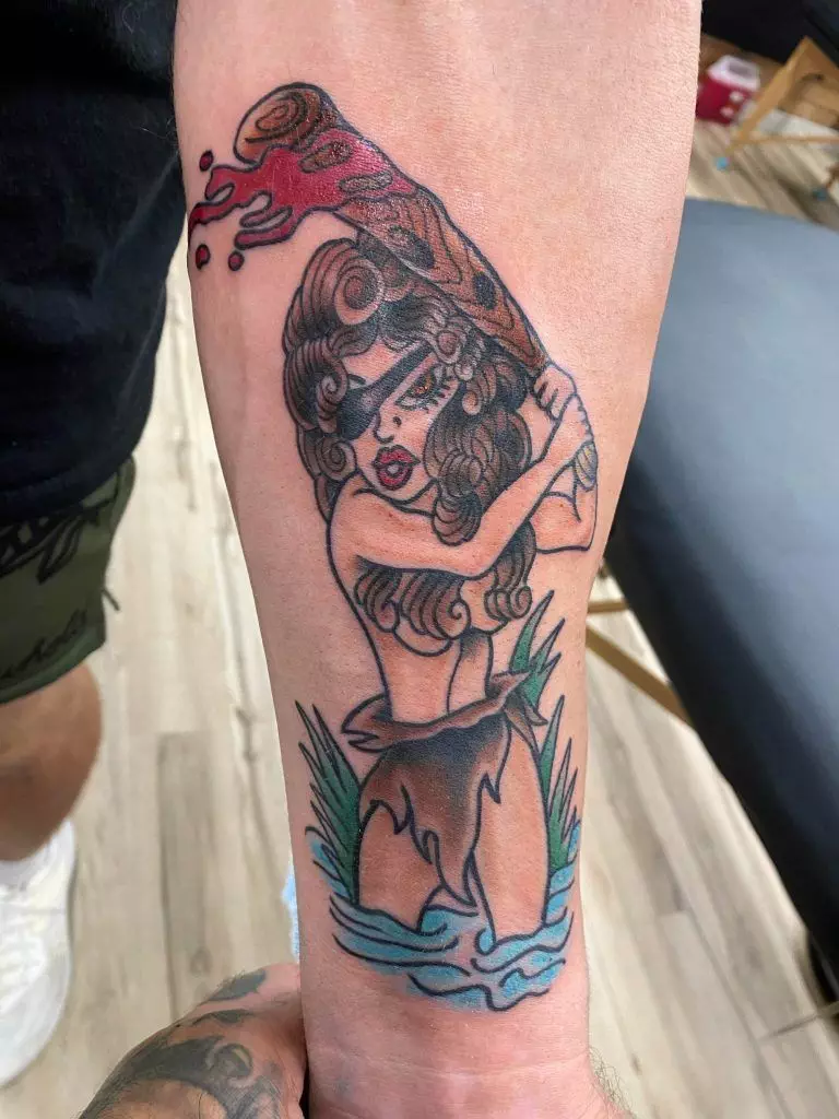 Forearm tattoo of a cartoon-style woman in a cavewoman outfit holding a bloodied baseball bat.