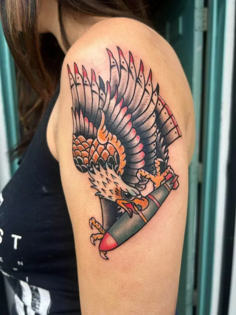 A colorful tattoo of an eagle holding a bomb on a person's upper arm.