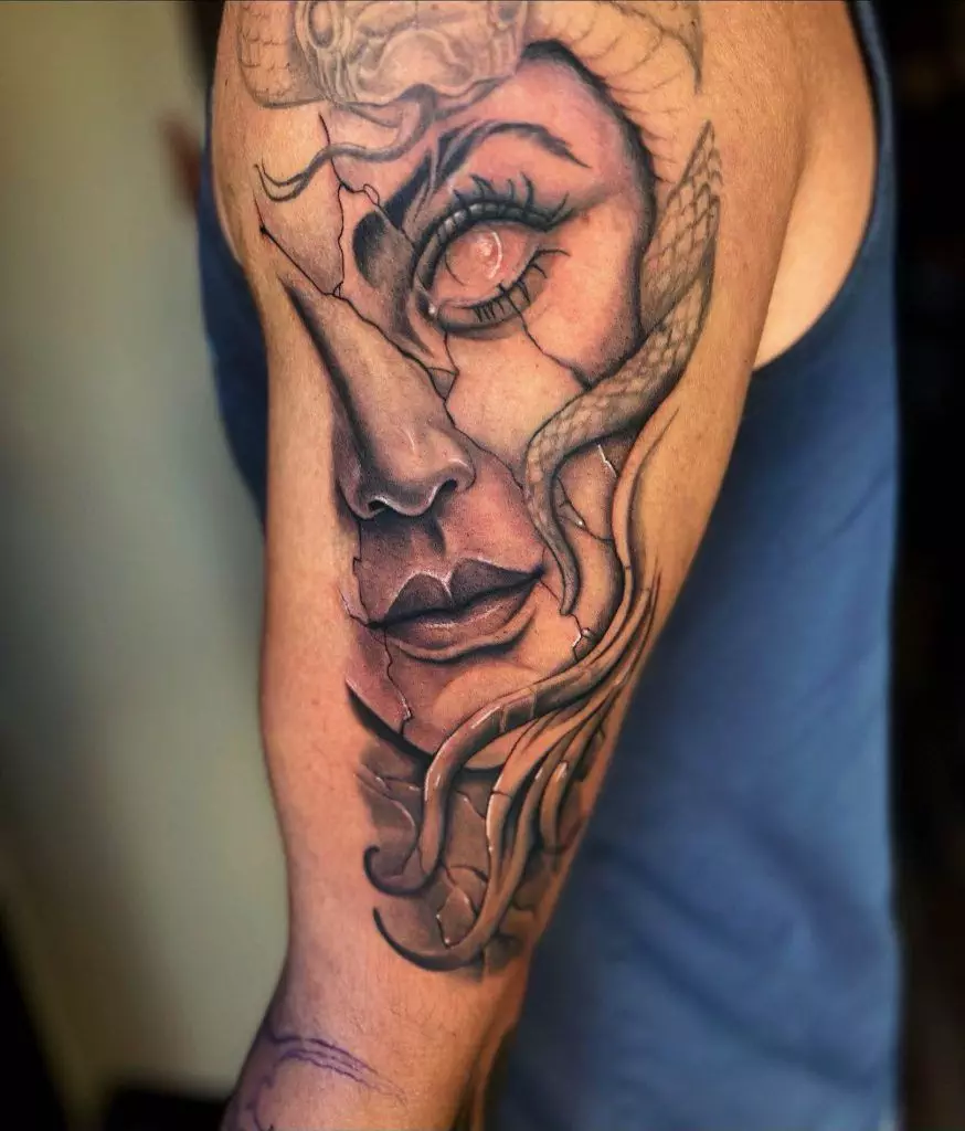 A detailed tattoo of a woman's face with abstract, cracked effects and snake motifs, inked on a person's upper arm.