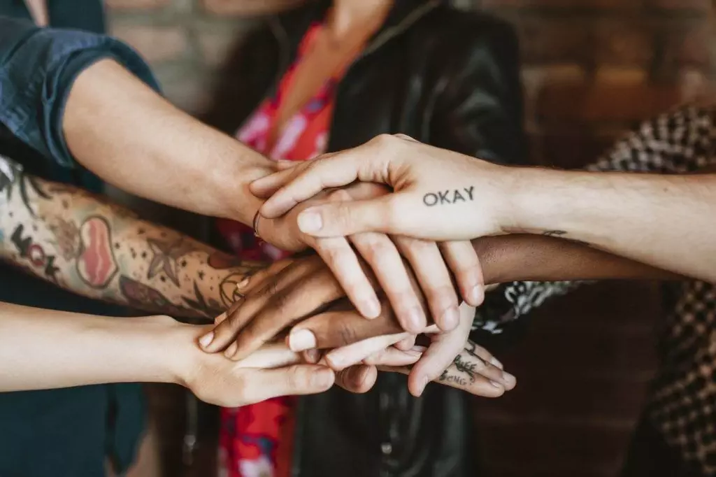 A group of people place their hands together in a stack. Some hands have tattoos, with one displaying the word 