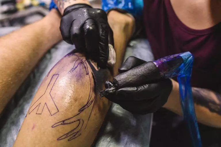 A tattoo artist wearing black gloves is using a tattoo machine to ink a design on a person's leg. The intricate tattoo, brought to life with vibrant pigment, includes lines and circular shapes.