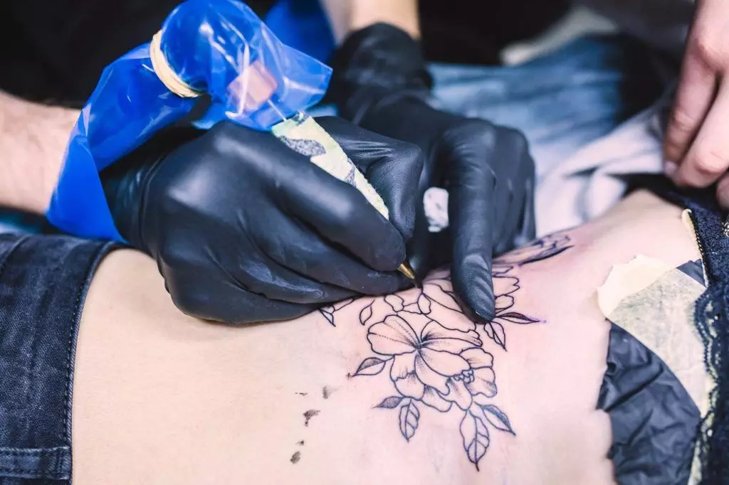 A tattoo artist, wearing black gloves, is meticulously tattooing a floral design on a person's lower torso during one of their focused tattoo sessions.