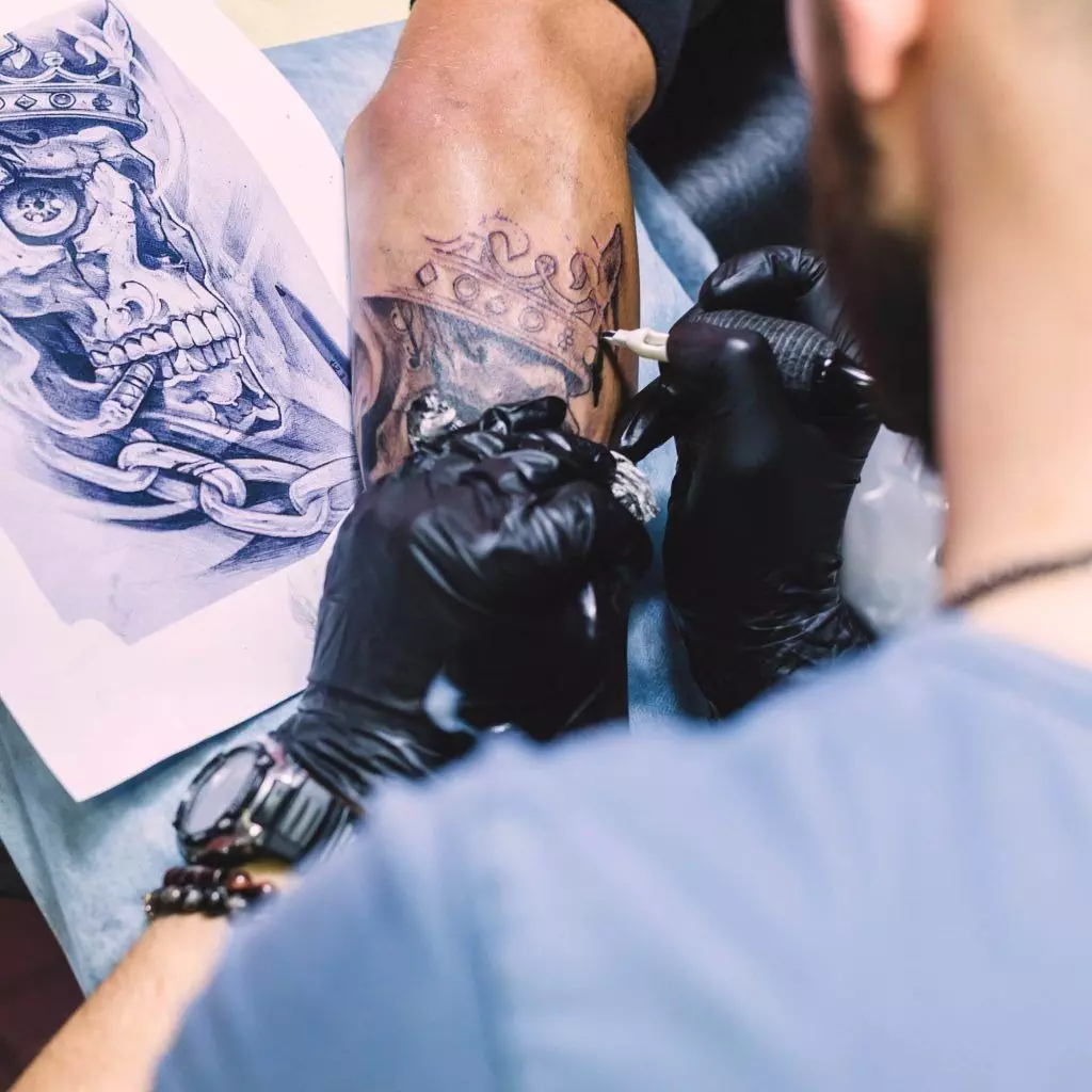 A tattoo artist in gloves works on a crown tattoo on a person's forearm, with a reference drawing of a skull and chains nearby, providing a step-by-step breakdown to ensure perfection during the tattoo session.