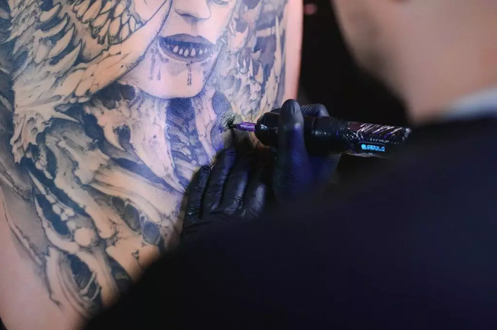 A tattoo artist with gloved hands injects vibrant pigment into a detailed, partially completed tattoo of a face and abstract designs on a person's back.