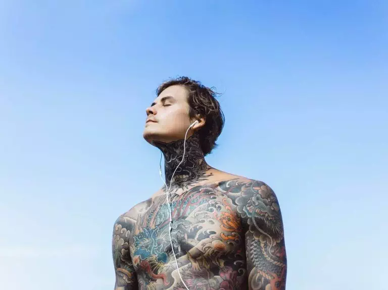 A tattooed man sporting a Dotwork Tattoo, with his eyes closed, enjoys the sunshine against a clear blue sky.