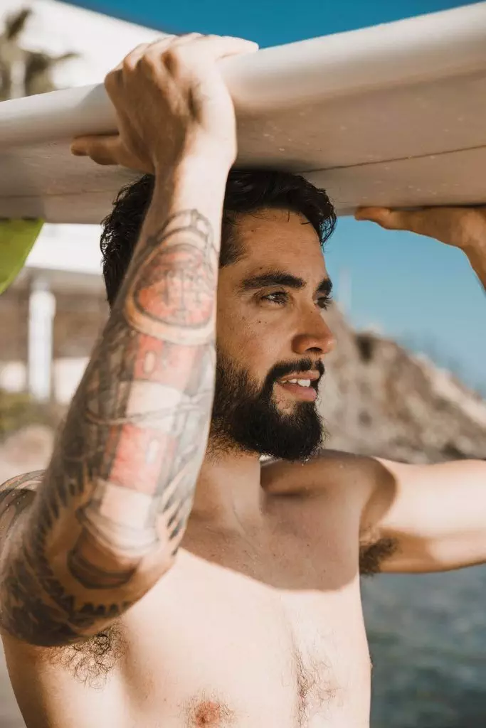 A shirtless man with a beard and arm tattoos holds a surfboard above his head on a sunny day at the beach, showcasing how to age with grace while preserving the beauty of an active lifestyle.