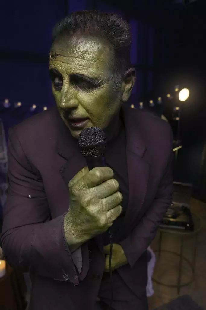 A person dressed as Frankenstein's monster, with green face paint and a suit, sings into a microphone in a dimly lit room, embodying one of the most iconic characters in the history of horror.