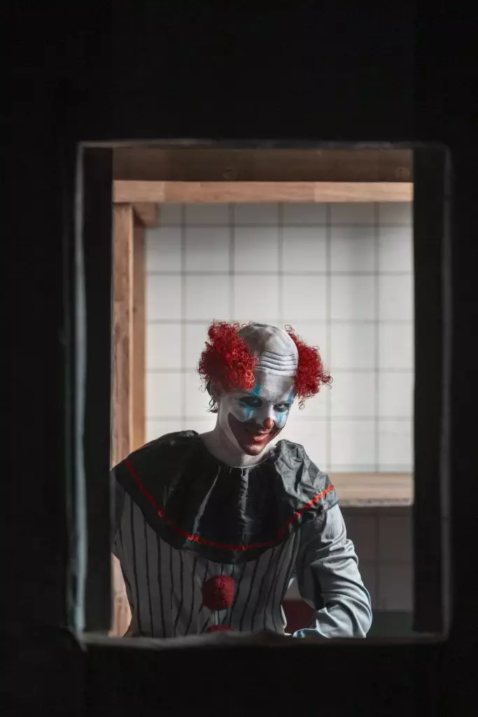 A person dressed as a clown with red curly hair and white face paint is seen through a window frame. The background shows a tiled wall and part of a wooden structure, evoking the eerie ambiance often associated with the history of horror.