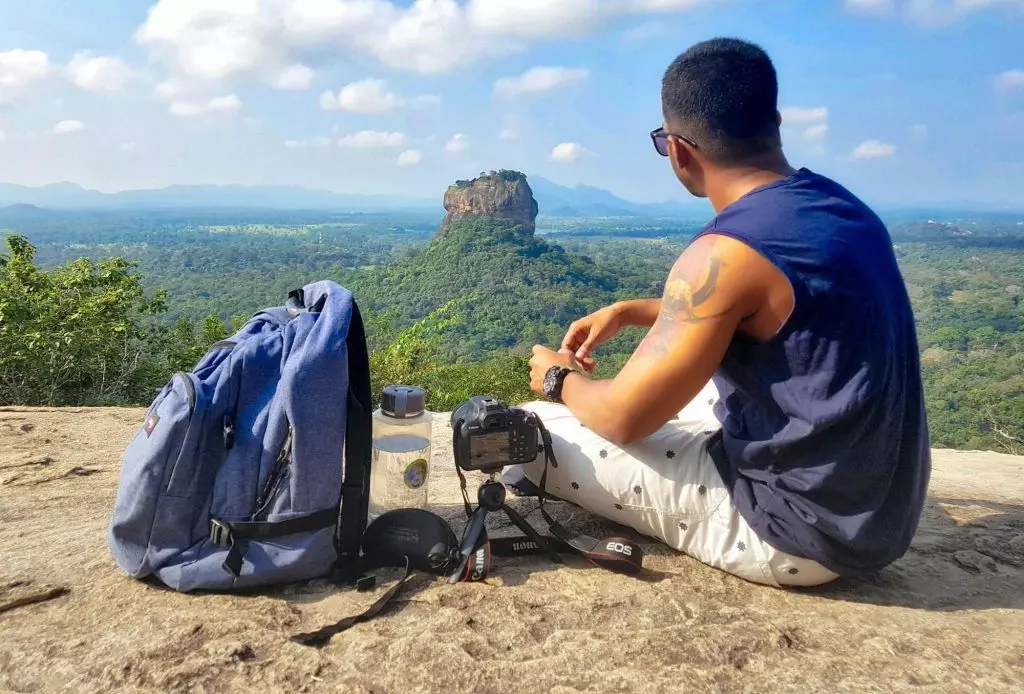 Man Sitting on Top of Gray Cliff Mountain Beside Backpack, Water Bottle, and Camera