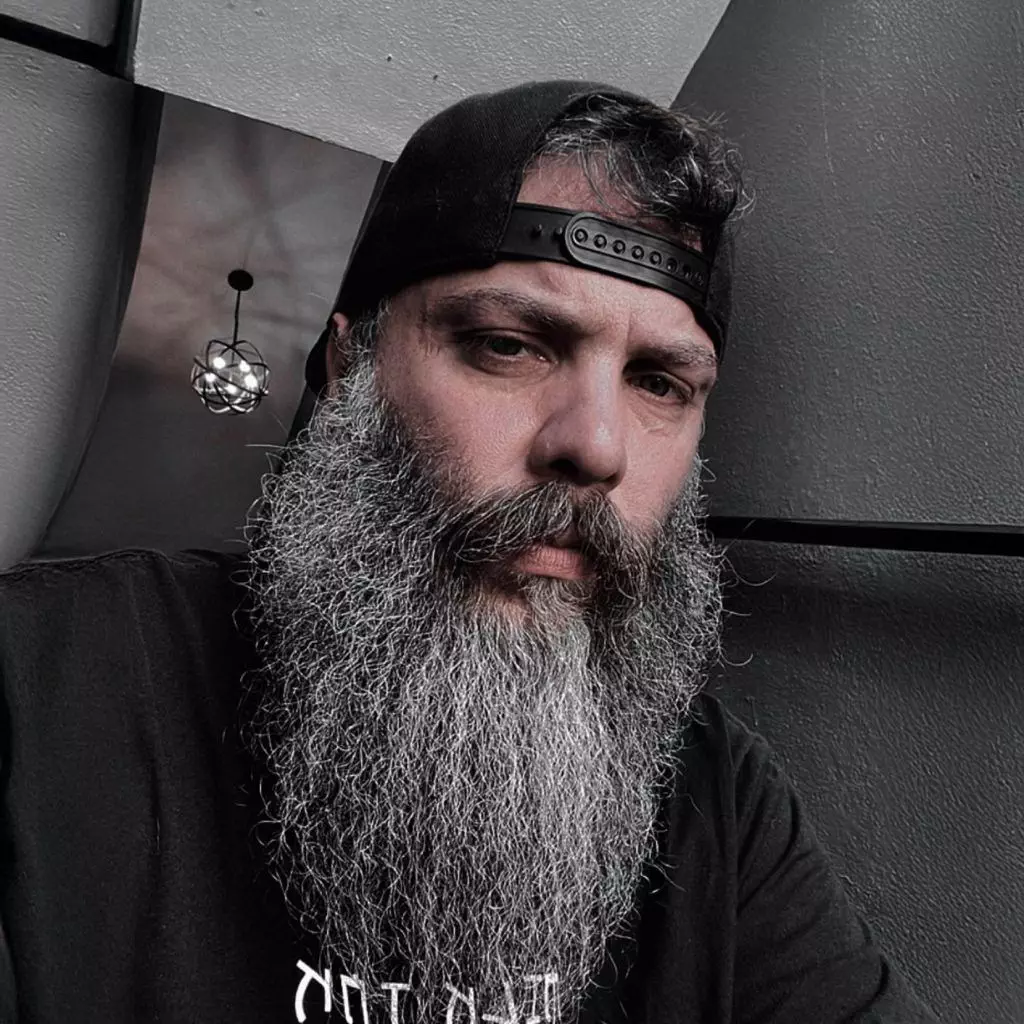 Man with a long gray beard wearing a backward black cap and a black shirt, sitting against a gray wall.