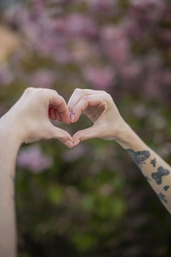 Hands form a heart shape with blooming flowers in the background, symbolizing love in a London park.